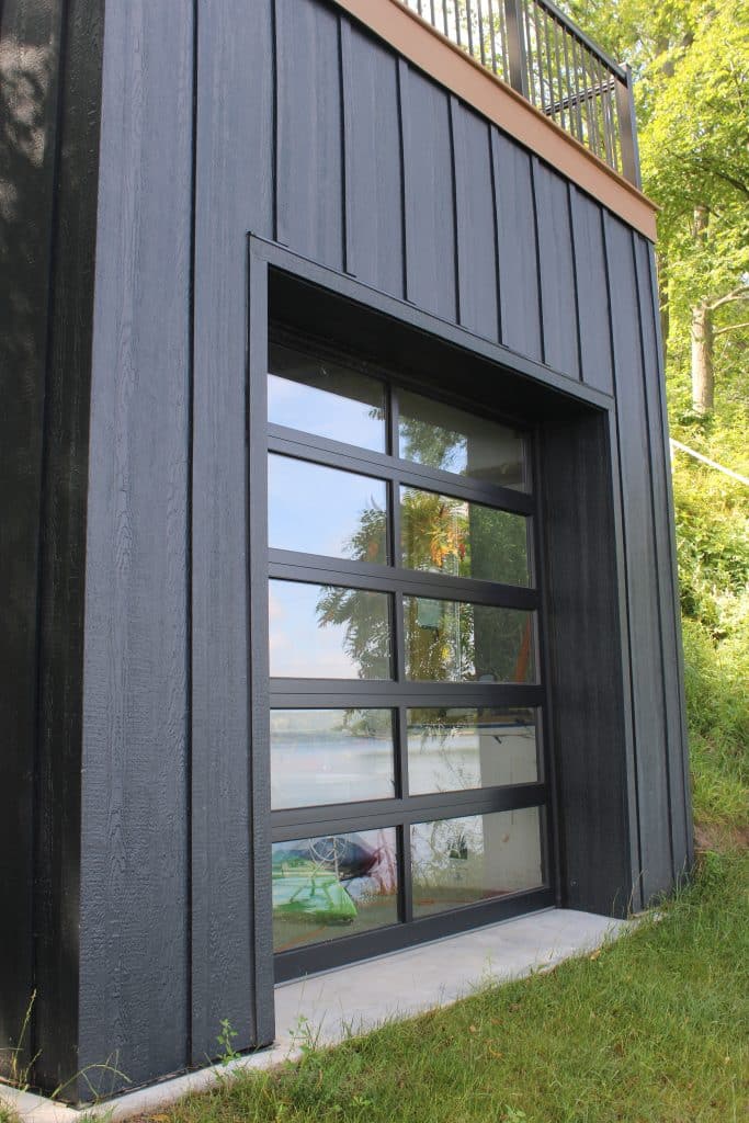 A modern black building with a large glass garage door, reflecting trees and a lake in the background. Surrounded by lush green grass and trees, it showcases sleek design—perfect for seamless garage door installation.