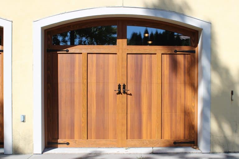 A wooden garage door with a natural finish, featuring vertical panels, black decorative hinges, and handles, set in a light-colored stucco wall with white arched trim—ideal for seamless garage door openers or professional garage door installation.