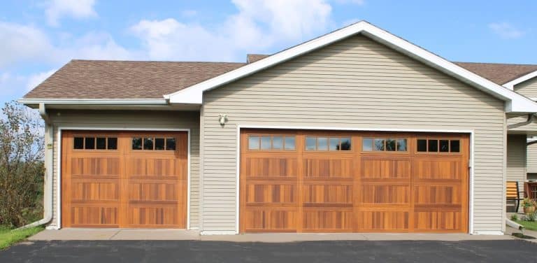 A beige house with two wooden garage doors, each with rectangular windows at the top. The driveway is paved and the sky is partly cloudy—perfect inspiration for anyone seeking a reliable Garage Door Company or garage door repair services.