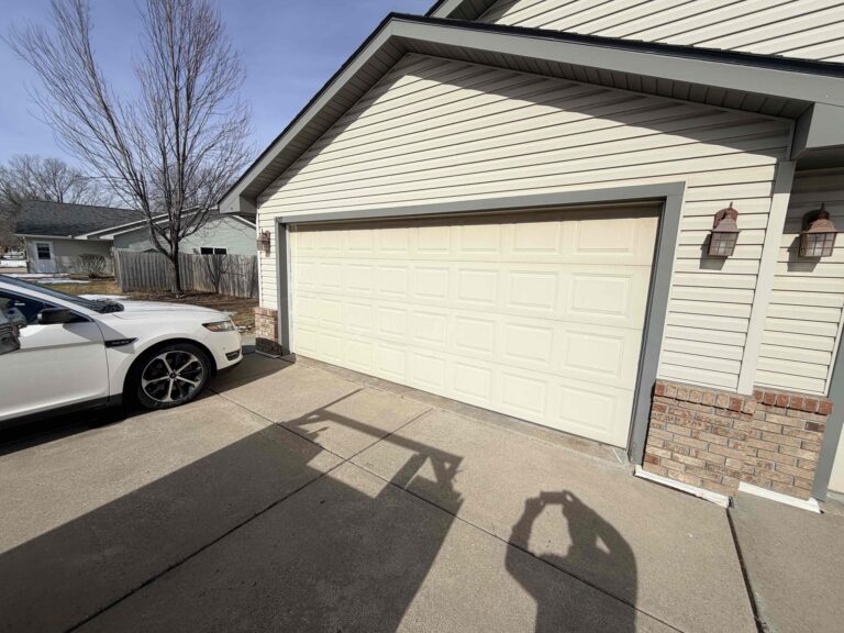 A closed beige garage door on a house with light siding and brick trim. A white car is parked in the driveway as a person's shadow appears taking a photo. Is it better to repair or replace your garage door? Two outdoor lights flank the garage.