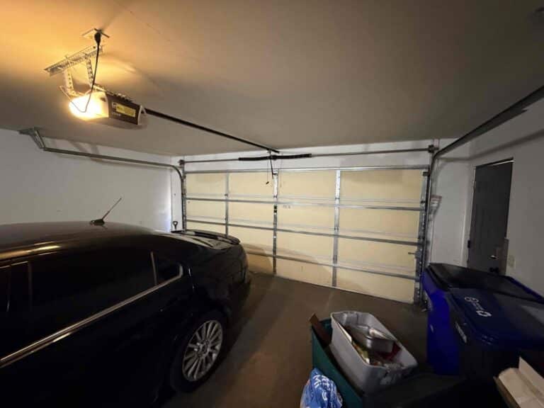 A dimly lit garage with a closed double garage door, a black car parked inside, blue recycling bins, and storage items along the right wall—prompting the question: Is it better to repair or replace your garage door? A ceiling-mounted opener is visible.