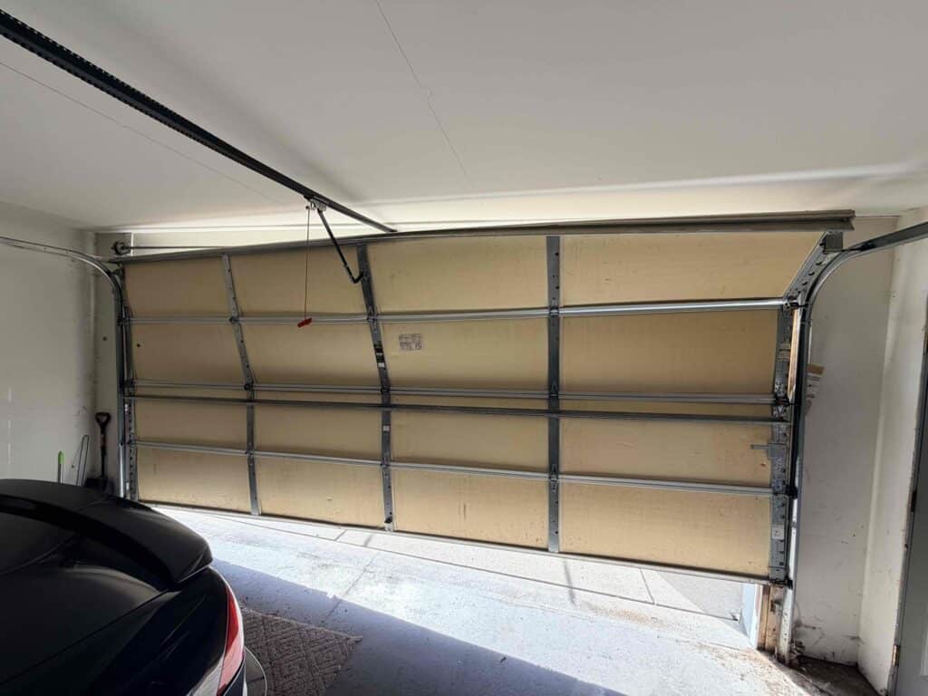 A partially open garage door appears bent and damaged in the middle, with daylight visible through gaps. The back of a black car is parked on the left side inside the garage—raising the question: Is it better to repair or replace your garage door?.