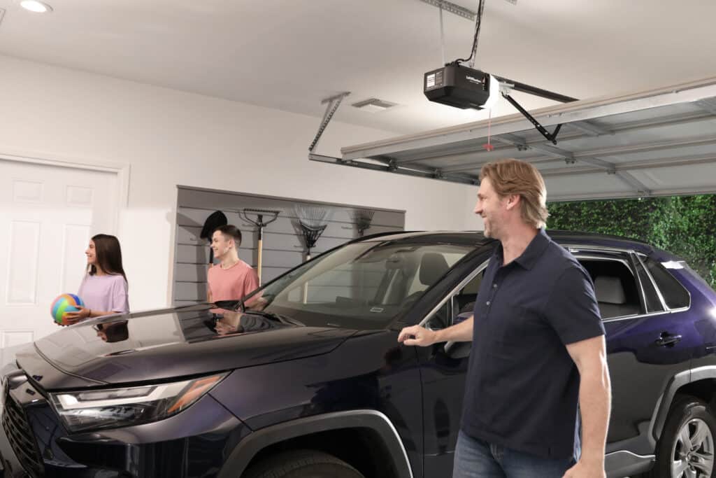 A man stands by a parked SUV in a garage, looking back at a smiling teenage boy and girl holding a ball near the door. The garage door is open, with garden tools mounted on the wall.