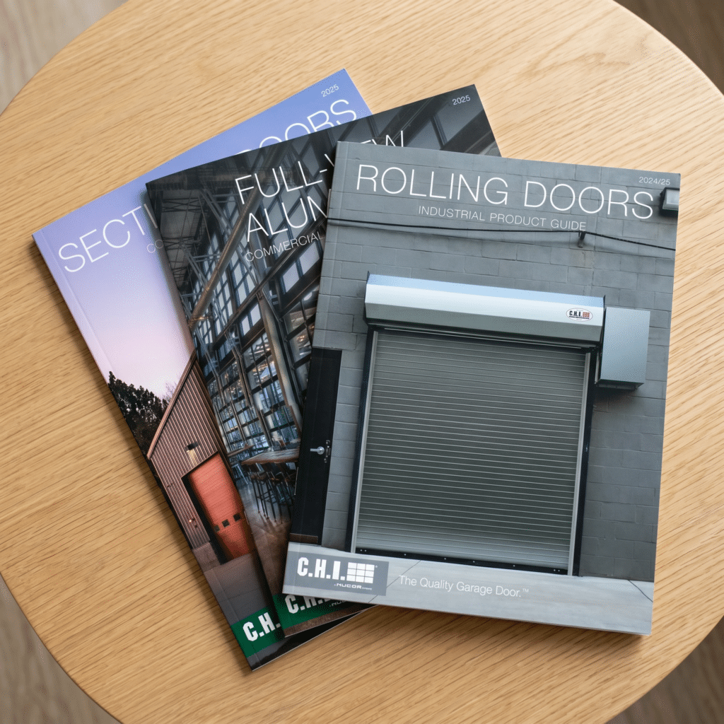 Three commercial door product catalogs are stacked on a wooden table. The top catalog features a photo of a gray rolling garage door on its cover and is titled Rolling Doors: Industrial Product Guide.