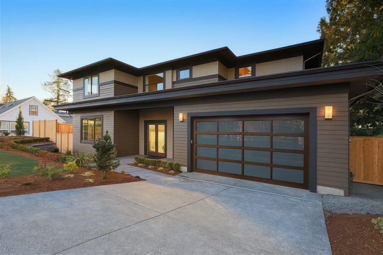 Modern two-story house with dark siding, large windows, and a glass-paneled garage door. The driveway is wide, and the front yard has fresh landscaping with mulch, shrubs, and a small tree. Evening lighting is on.