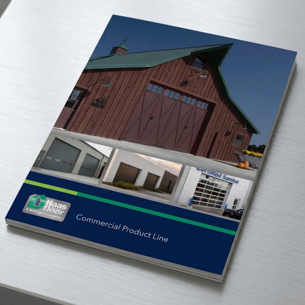 A brochure on a metal surface features photos of different commercial garage doors and buildings. The cover includes the Haas Door logo and the title “Commercial Product Line.”.