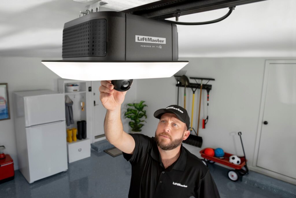 A man in a LiftMaster uniform adjusts a camera on a LiftMaster garage door opener in a clean, organized garage with tools, toys, and a refrigerator in the background.