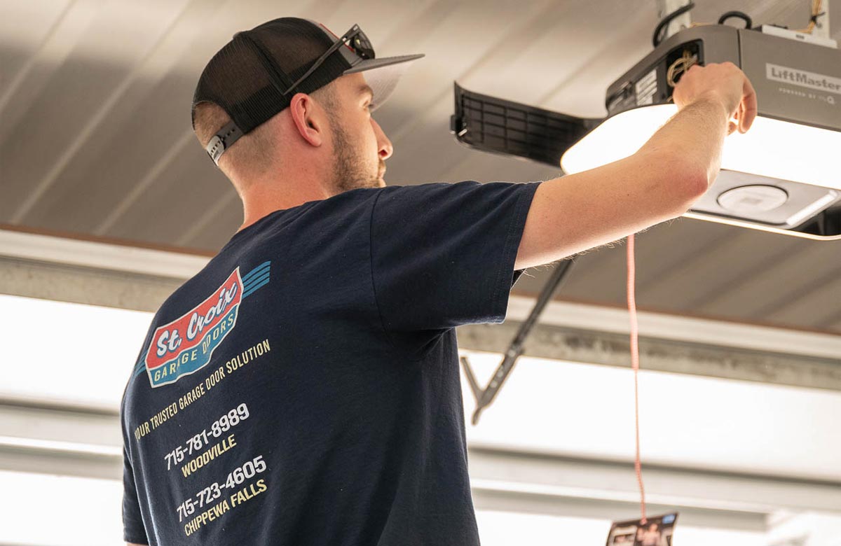 A man wearing a cap and a blue t-shirt is adjusting a garage door opener inside a garage. The back of his shirt lists business contact information.